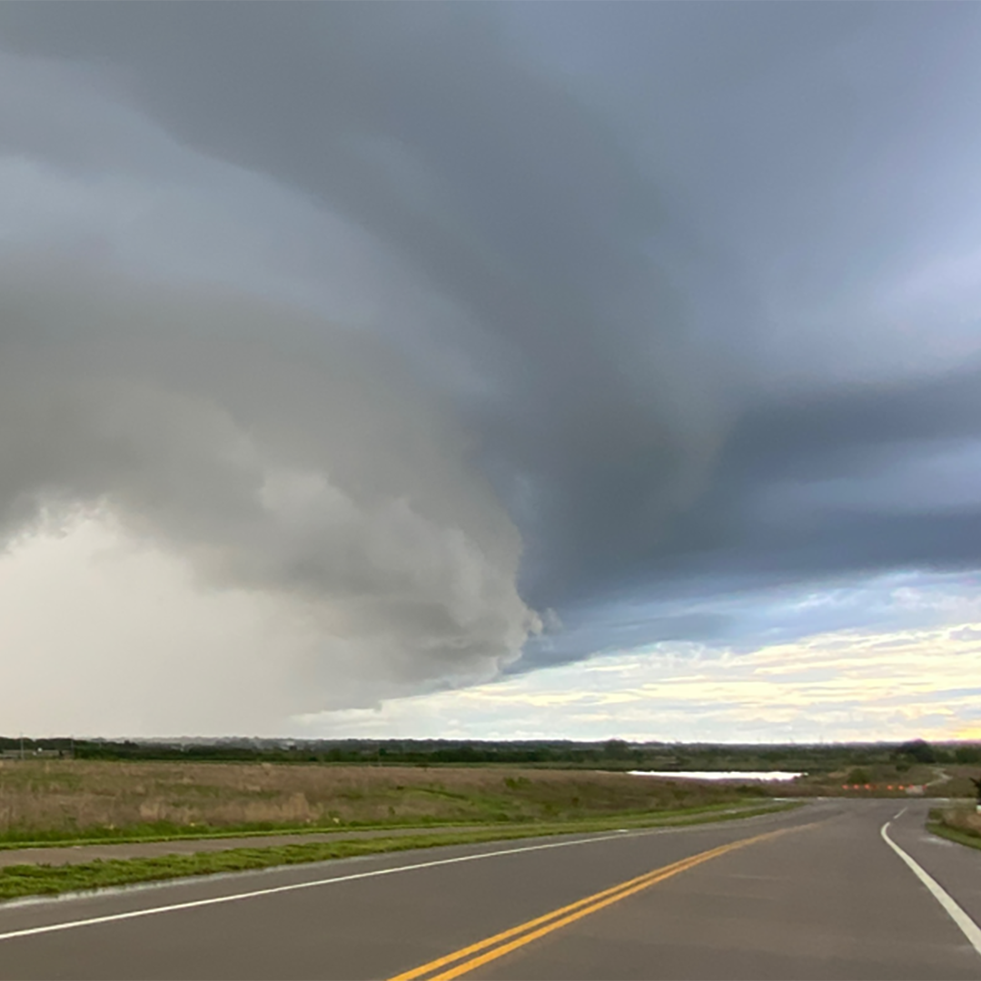 Large, dark storm clouds over a winding rural highway, with grassy fields and distant horizon beneath the approaching weather.